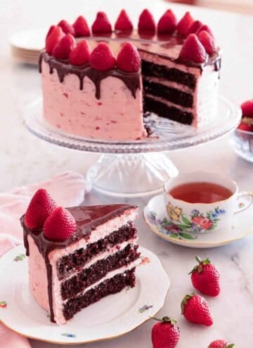 A slice of chocolate strawberry cake on a plate in front of a cake stand with the rest of the cake.