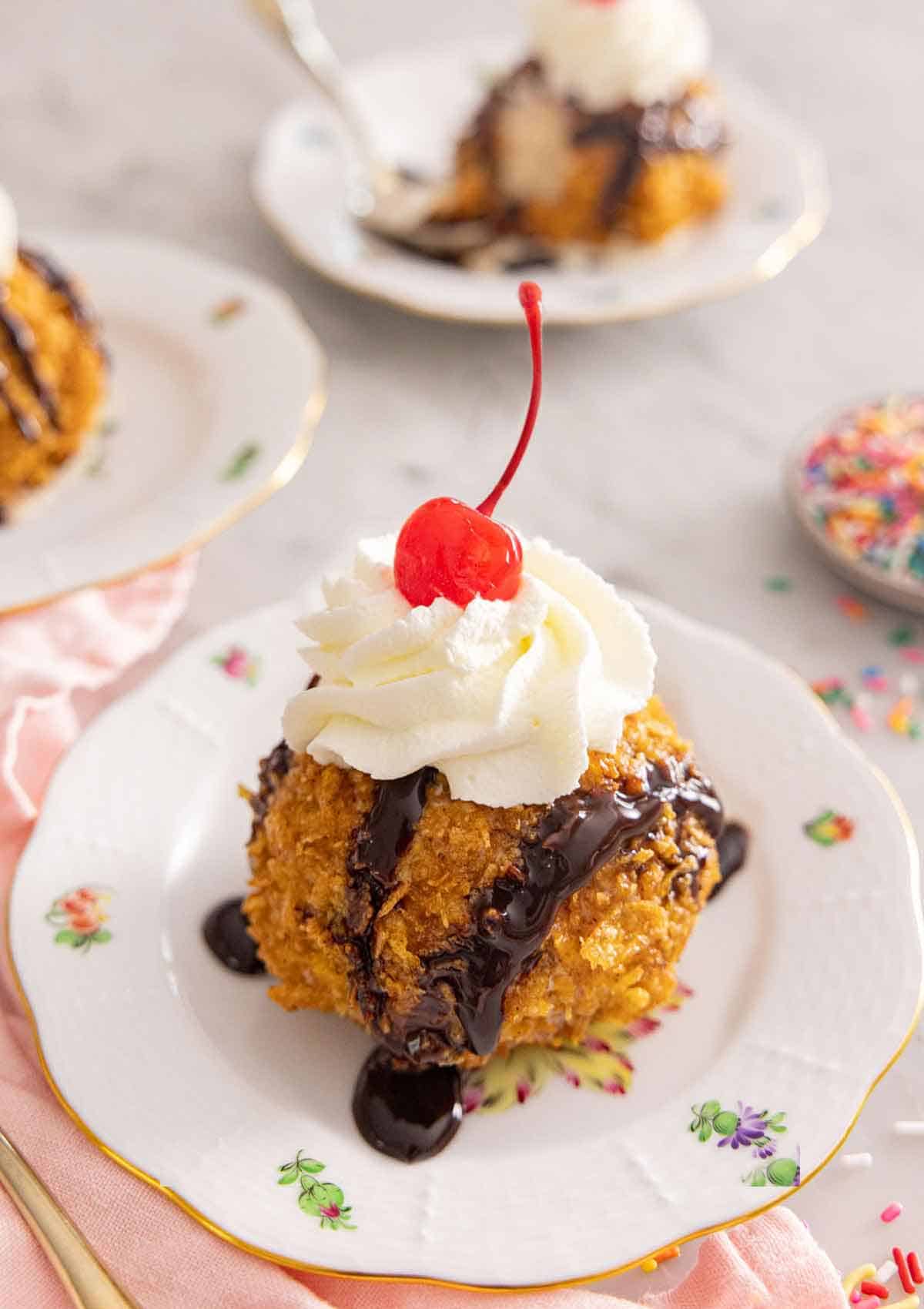 A plate with a ball of fried ice cream with chocolate drizzle and whipped cream on top with a cherry.