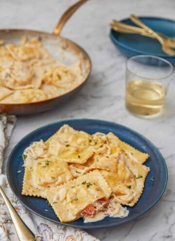 A plate of lobster ravioli by a glass of wine and a skillet with pasta.
