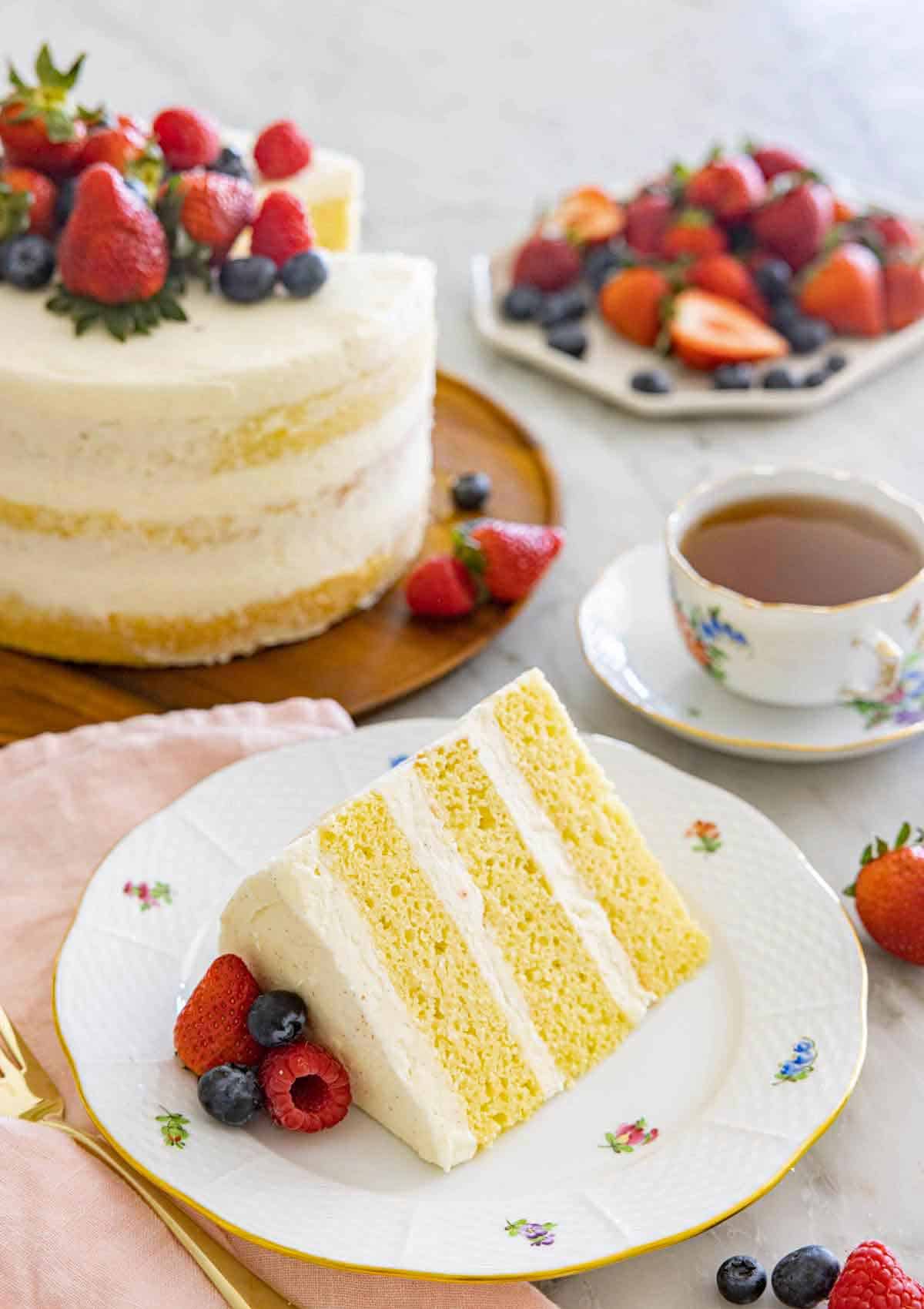 A plate with a slice of three layered naked cake by a mug of tea and the rest of the cake.