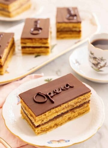 A serving of opera cake on a plate by a cup of coffee with more cakes in the background.