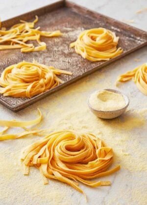 Multiple bundles of pasta dough on a sheet pan with one in the foreground with a small bowl of semolina flour.