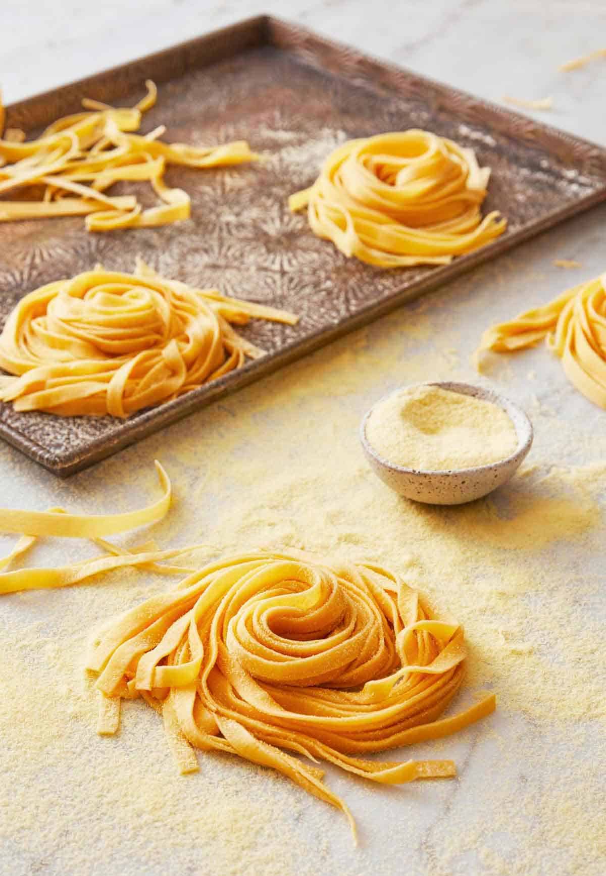 Multiple bundles of pasta dough on a sheet pan with one in the foreground with a small bowl of semolina flour.