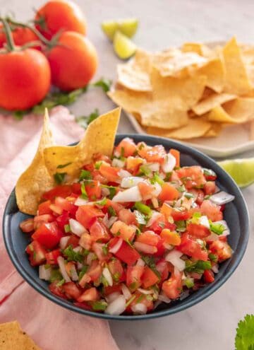 A bowl of pico de gallo with two tortilla chips in the bowl and more in the background.