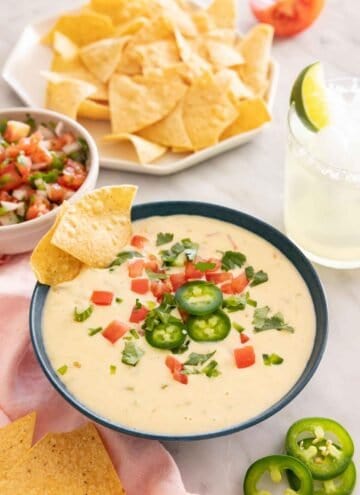 A bowl of queso dip with various toppings with a plate of chips and pico de gallo in the background.