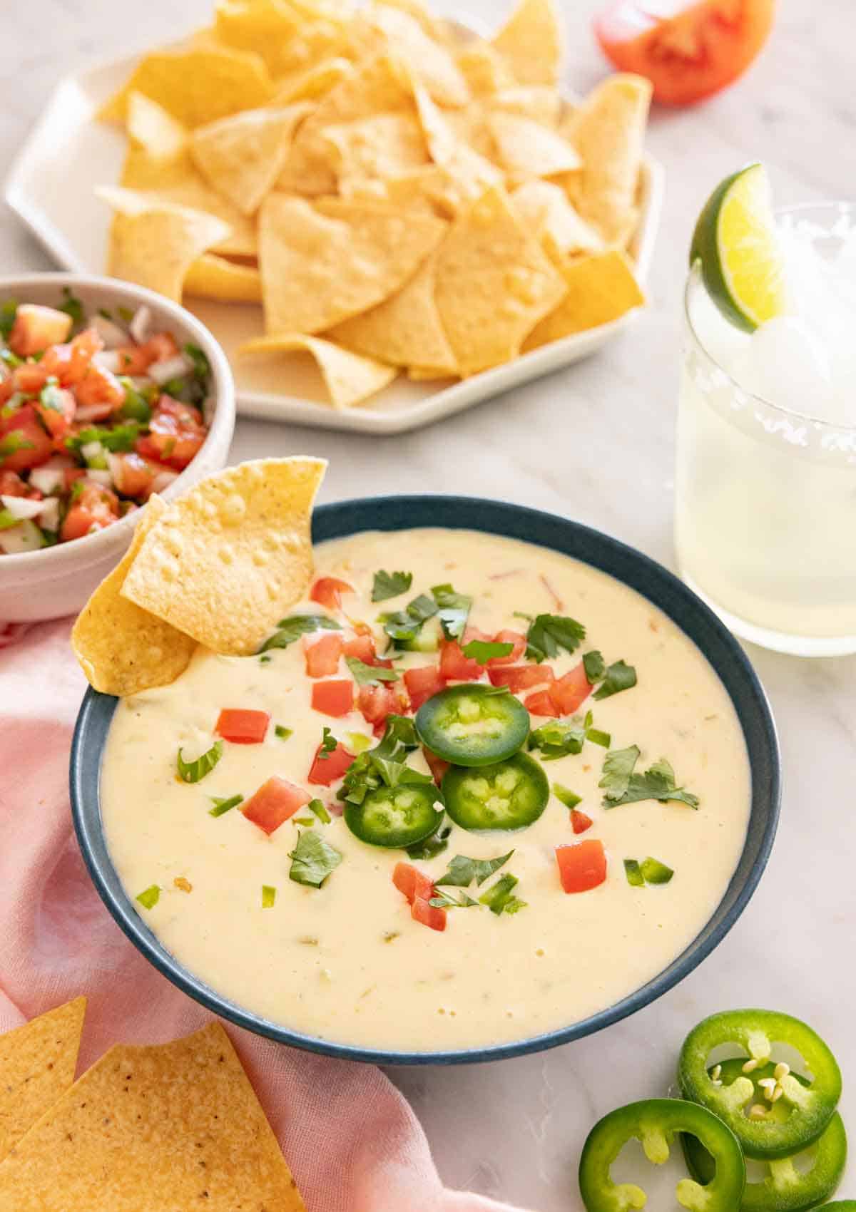 A bowl of queso dip with various toppings with a plate of chips and pico de gallo in the background.