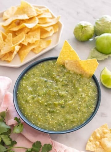 A bowl of salsa verde with two tortilla chips in the bowl with more ingredients scattered around it.