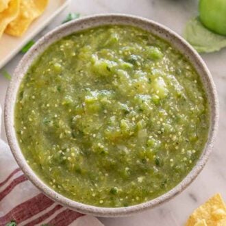 Overhead view of a bowl of salsa verde with some limes, chips, and tomatillos around it.