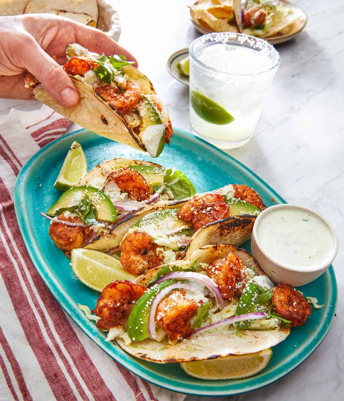 A hand lifting a shrimp taco off a platter with three more shrimp tacos with a small bowl of dip.