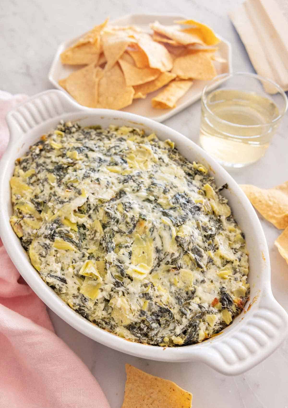 A white baking dish of spinach artichoke dip with a plate of chips and glass of wine behind it.