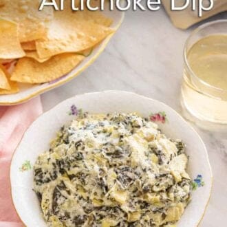 Pinterest graphic of a bowl of spinach artichoke dip. A plate of chips and glass of wine is beside the dip.