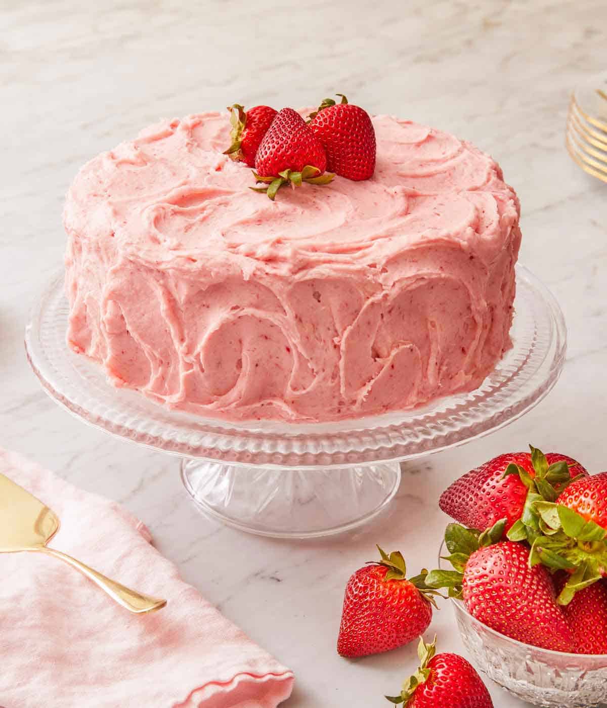 A strawberry cake on a cake stand with fresh strawberries on top and on the side.