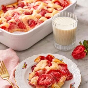 A plate with a serving of strawberry cobbler in front of a glass of milk and baking dish.