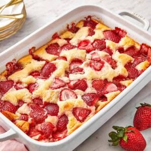A baking dish with a strawberry cobbler with two fresh strawberries on the side and a stack of plates.