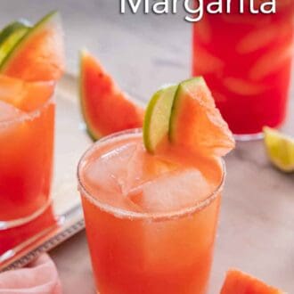 Pinterest graphic of a glass of watermelon margarita with two in the background. Cut watermelon and lime placed on the salted rim.