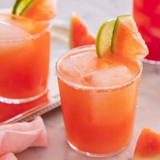 Glasses of watermelon margaritas, with one on a tray, with cut watermelon and limes in the drink and scattered around.