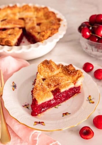 A slice of cherry pie on a plate in front of the rest of the pie in the background.