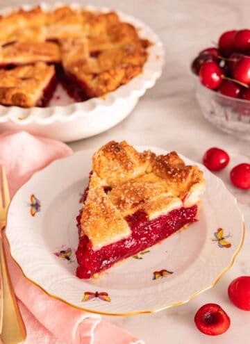 A slice of cherry pie on a plate in front of the rest of the pie in the background.