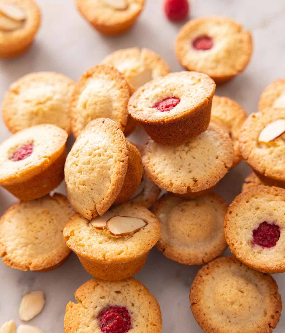 Multiple financiers in a pile, some plain, some with a raspberry on top, and some with sliced almonds on top.