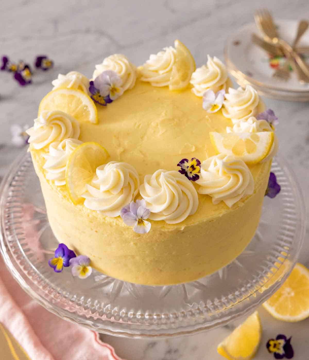 Overhead angled view of a lemon cake on a cake stand with edible purple flowers scattered around the cake and on the frosting.