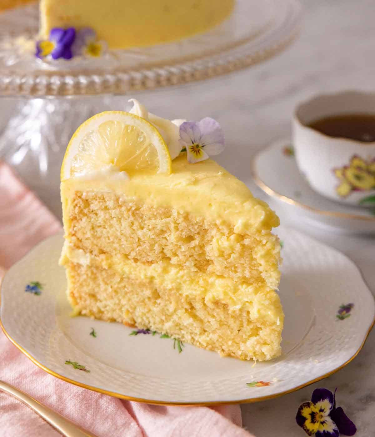 A slice of lemon cake with a slice of lemon on top with an edible flower for garnish.