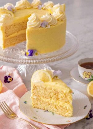 A slice of lemon cake on a plate in front of a cake stand holding the rest of the cake.