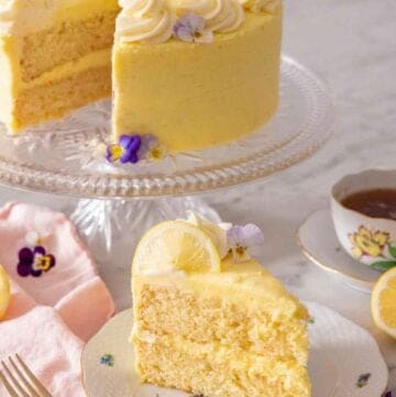 A slice of lemon cake on a plate in front of a cake stand holding the rest of the cake.