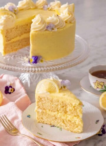 A slice of lemon cake on a plate in front of a cake stand holding the rest of the cake.