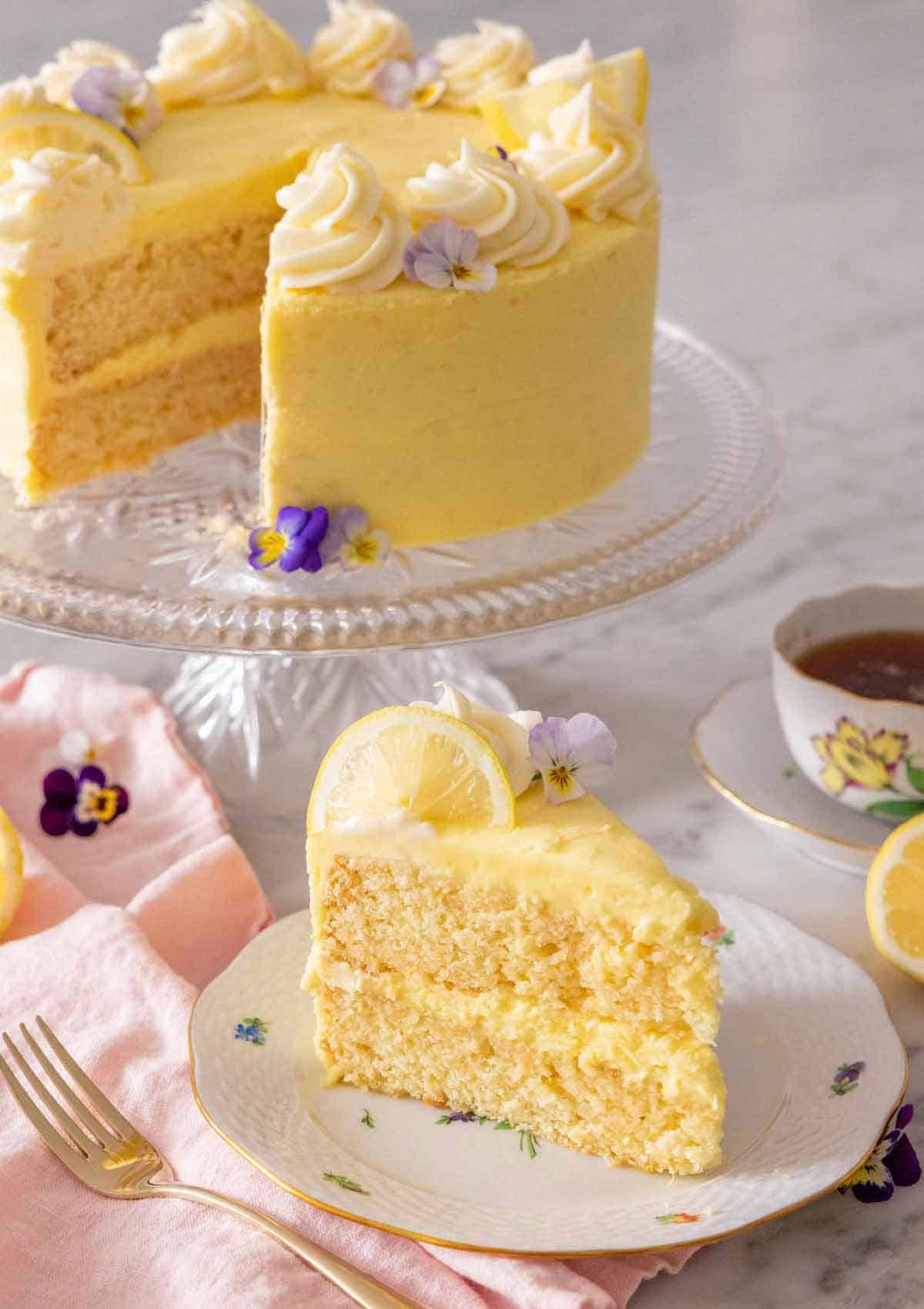 A slice of lemon cake on a plate in front of a cake stand holding the rest of the cake.