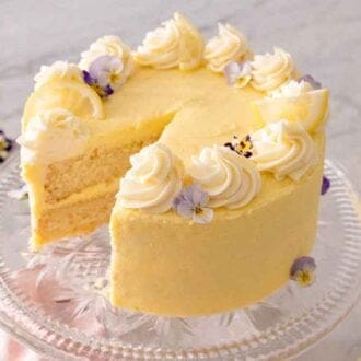 A lemon cake with a slice cut out on top of a cake stand with cut lemons and purple flowers scattered around on the counter.