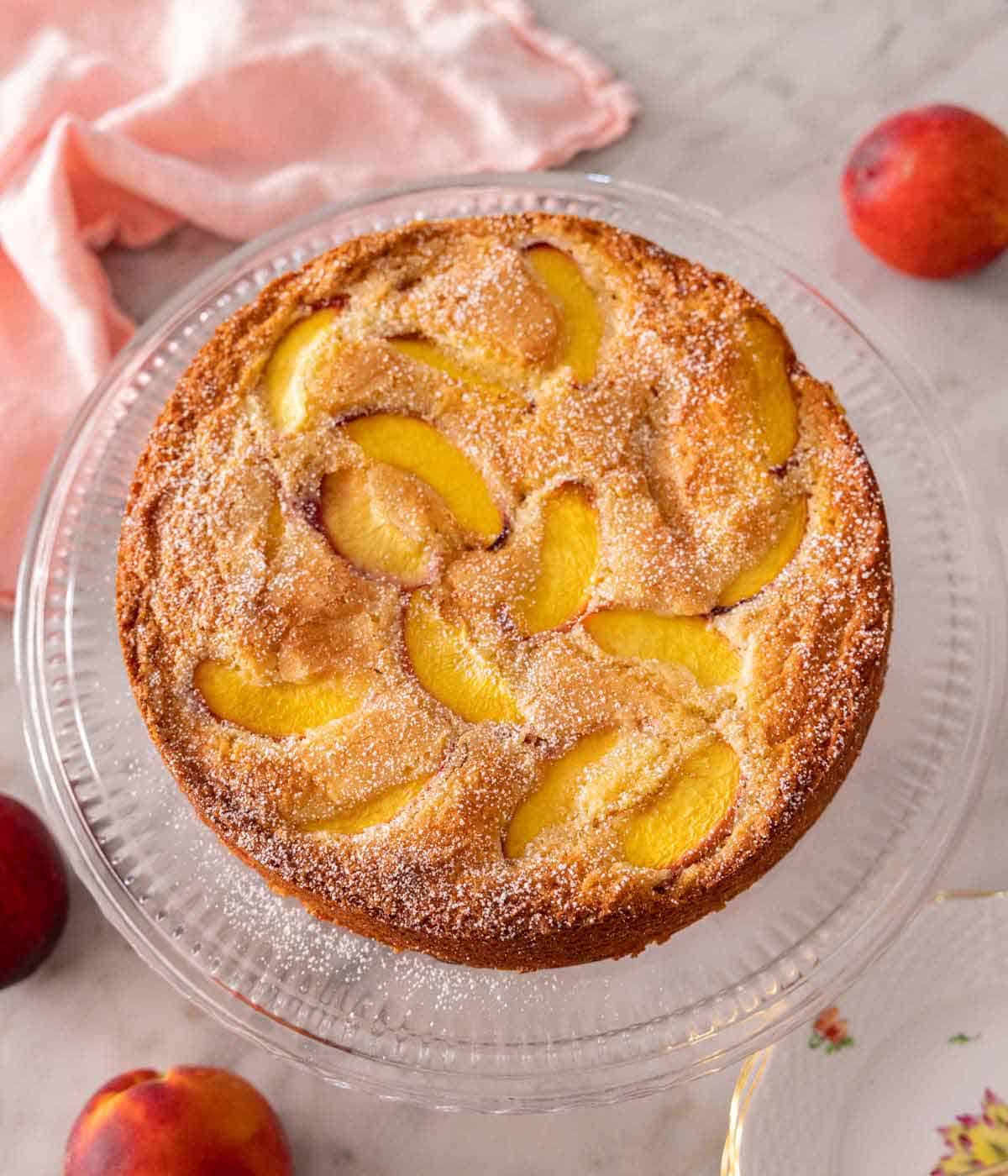 Overhead view of a peach cake on a cake stand with fresh peaches scattered around.