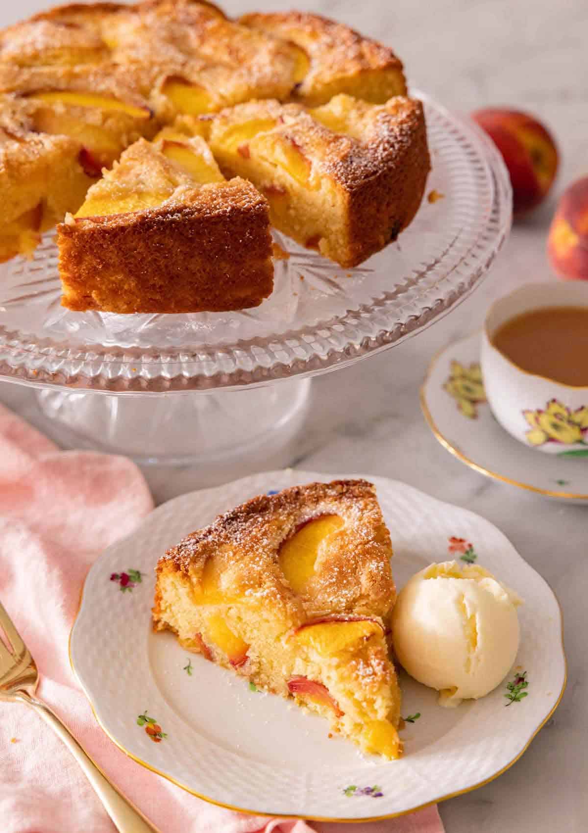 A slice of peach cake with a scoop of ice cream by a cake stand with the rest of the peach cake.