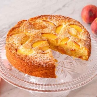 A cake stand with a peach cake on top with a slice cut out and plated in the background with two fresh peaches on the side.