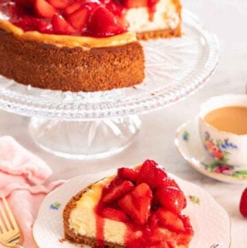 A slice of strawberry cheesecake with the syrup dripping off it, in front of a cake stand with the rest of the cheesecake.