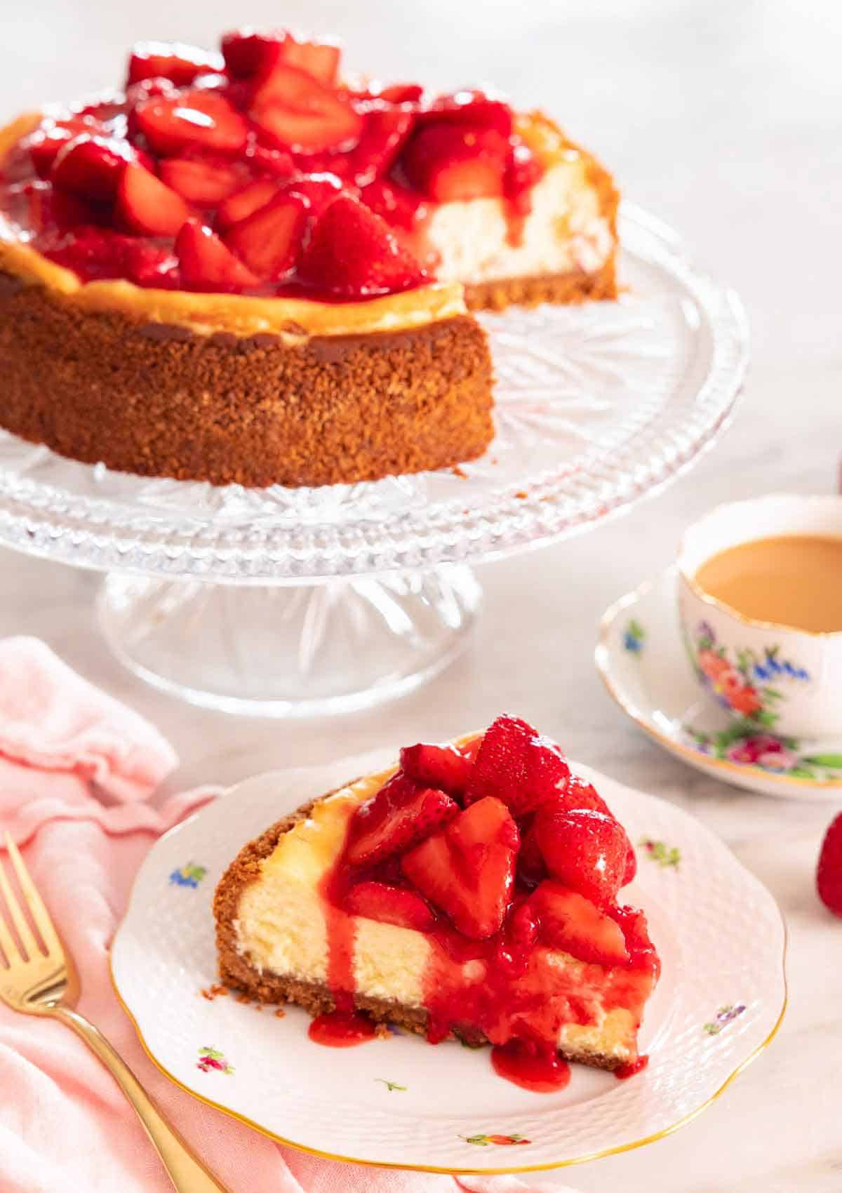 A slice of strawberry cheesecake with the syrup dripping off it, in front of a cake stand with the rest of the cheesecake.