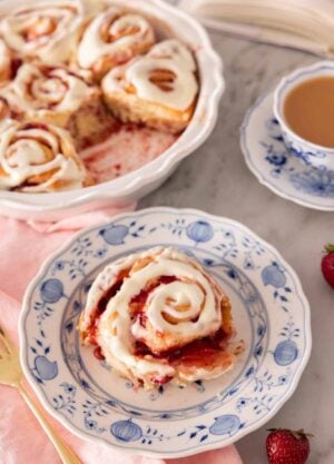 A plate with a serving of strawberry roll with cream cheese icing on top, by a baking dish with the rest of the rolls.