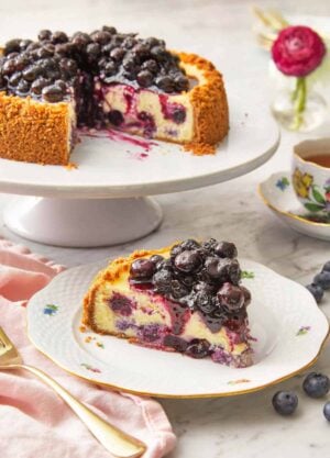 A slice of blueberry cheesecake on a plate in front of a cake stand with the rest of the cheesecake.