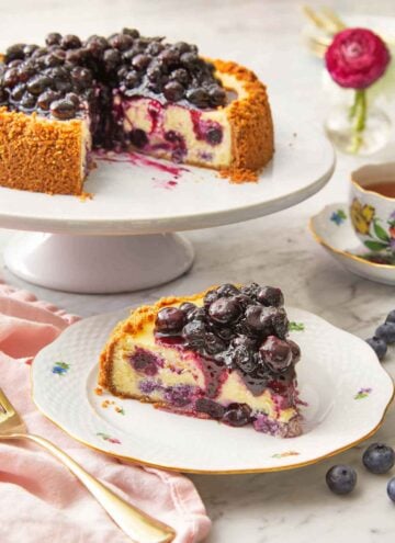 A slice of blueberry cheesecake on a plate in front of a cake stand with the rest of the cheesecake.