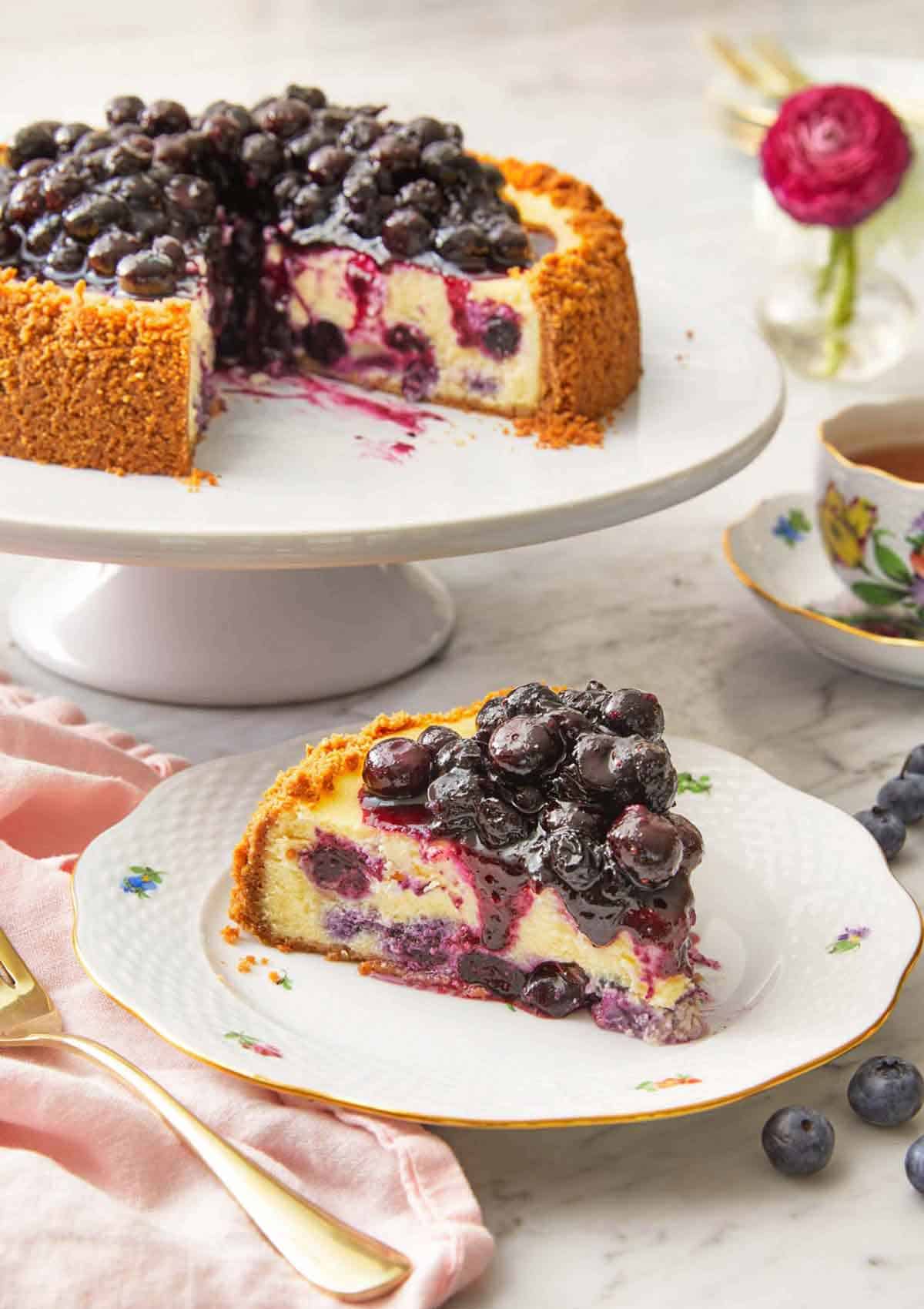 A slice of blueberry cheesecake on a plate in front of a cake stand with the rest of the cheesecake.