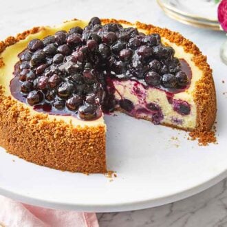 A white cake stand with a blueberry cheesecake with a slice cut out.