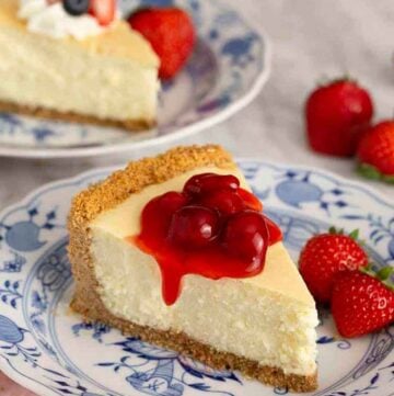 A slice of cheesecake with berry sauce on top with another slice in the background and strawberries scattered around.