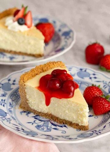 A slice of cheesecake with berry sauce on top with another slice in the background and strawberries scattered around.