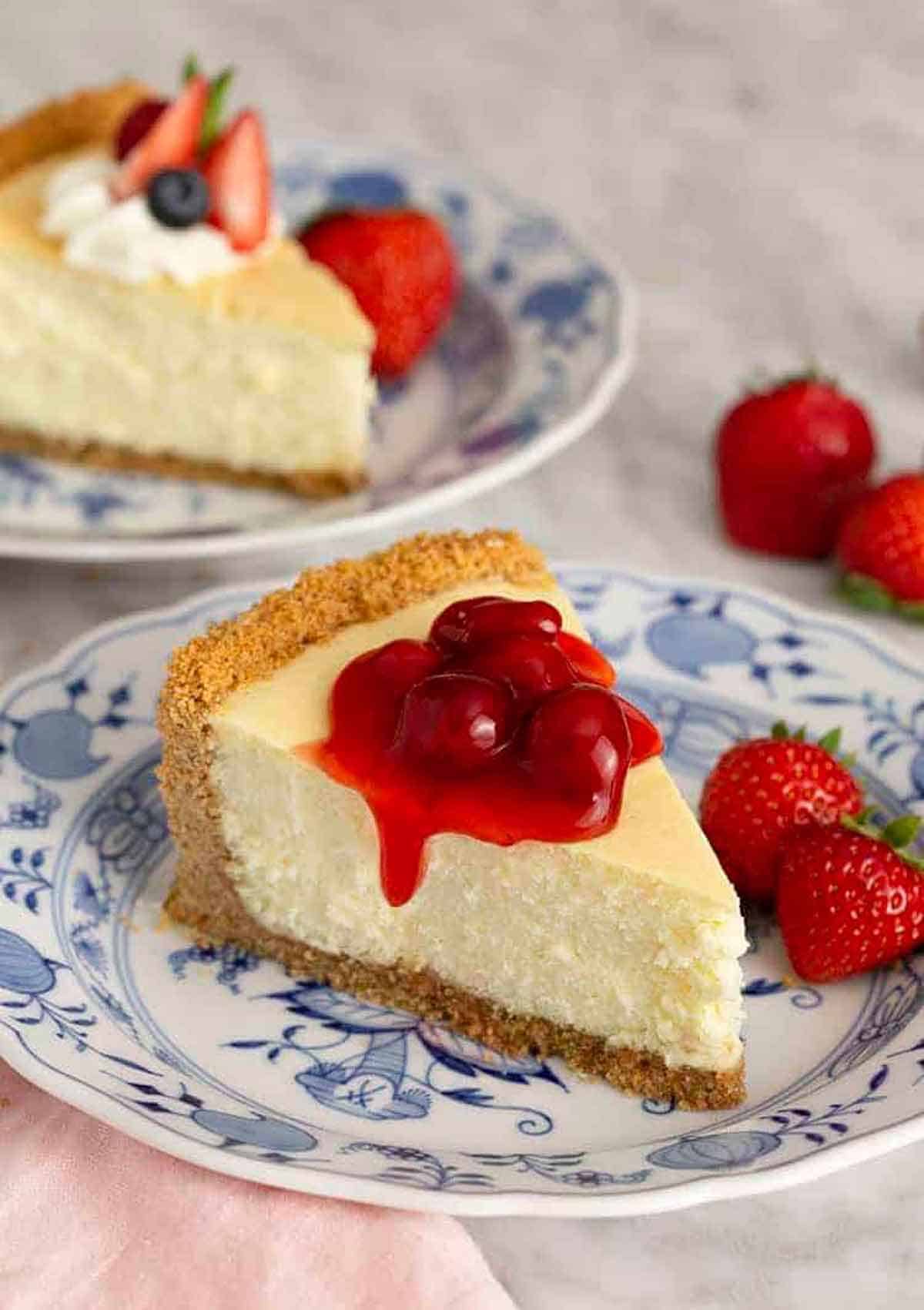 A slice of cheesecake with berry sauce on top with another slice in the background and strawberries scattered around.
