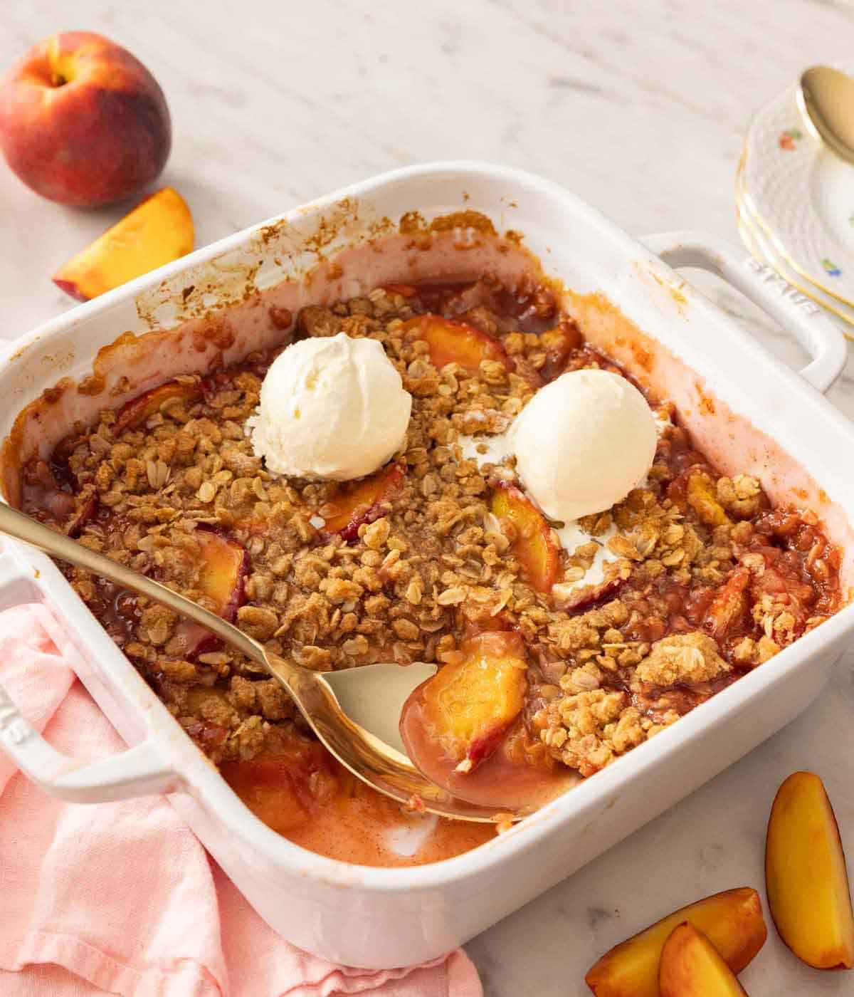 A square baking dish with peach crisp with a spoon tucked in with two scoops of vanilla ice cream on top.