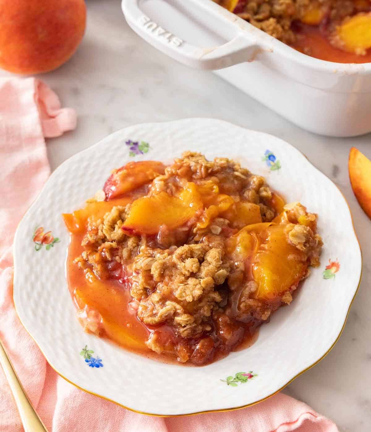 A plate of peach crisp with large chunks of peaches under the crispy oats on top of a pink napkin by a baking dish.