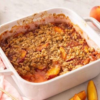 A white baking dish of peach crisp with a portion removed. Fresh peaches and slices scattered around.