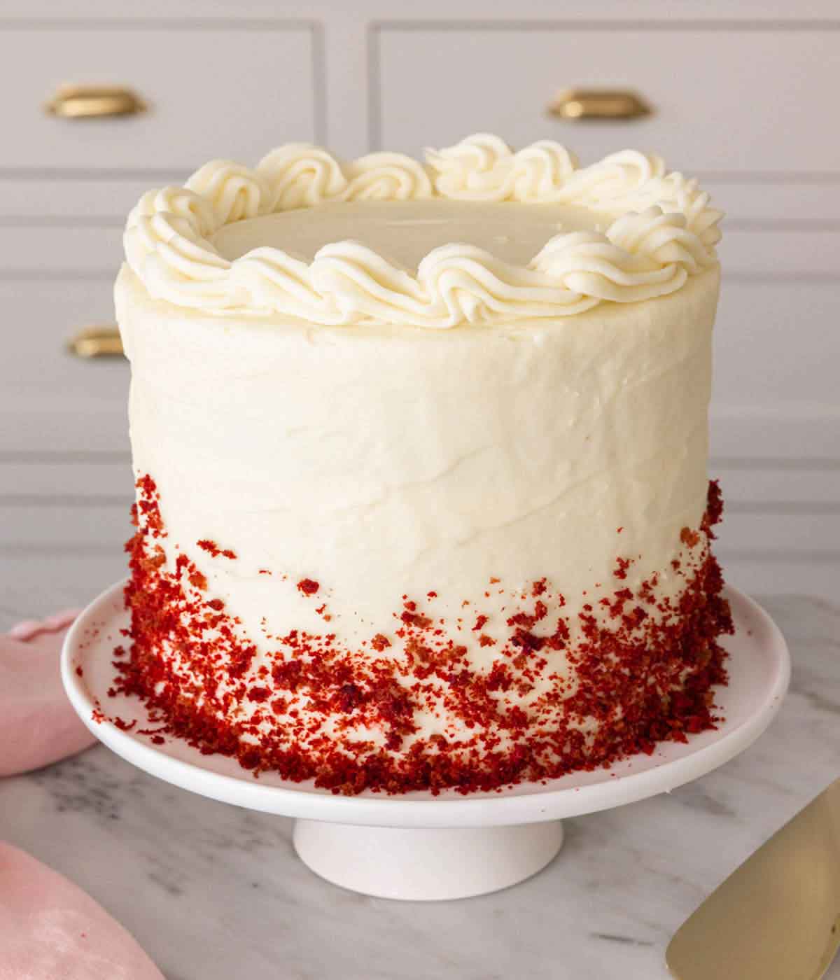 A red velvet cake on a cake stand with crumbled cake on the frosting.