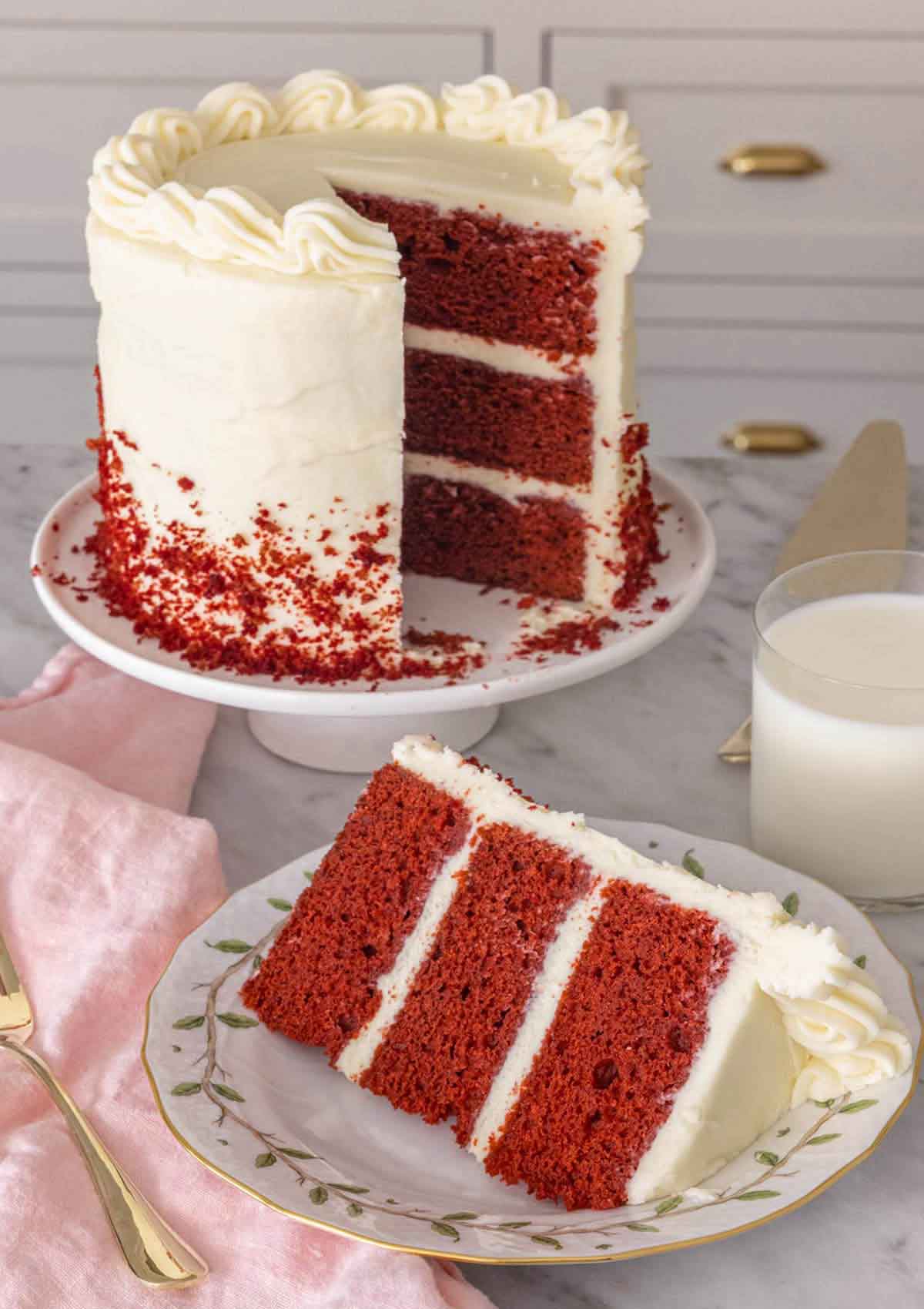 A cake stand with a slice of red velvet cake cut out and placed on a plate in front.