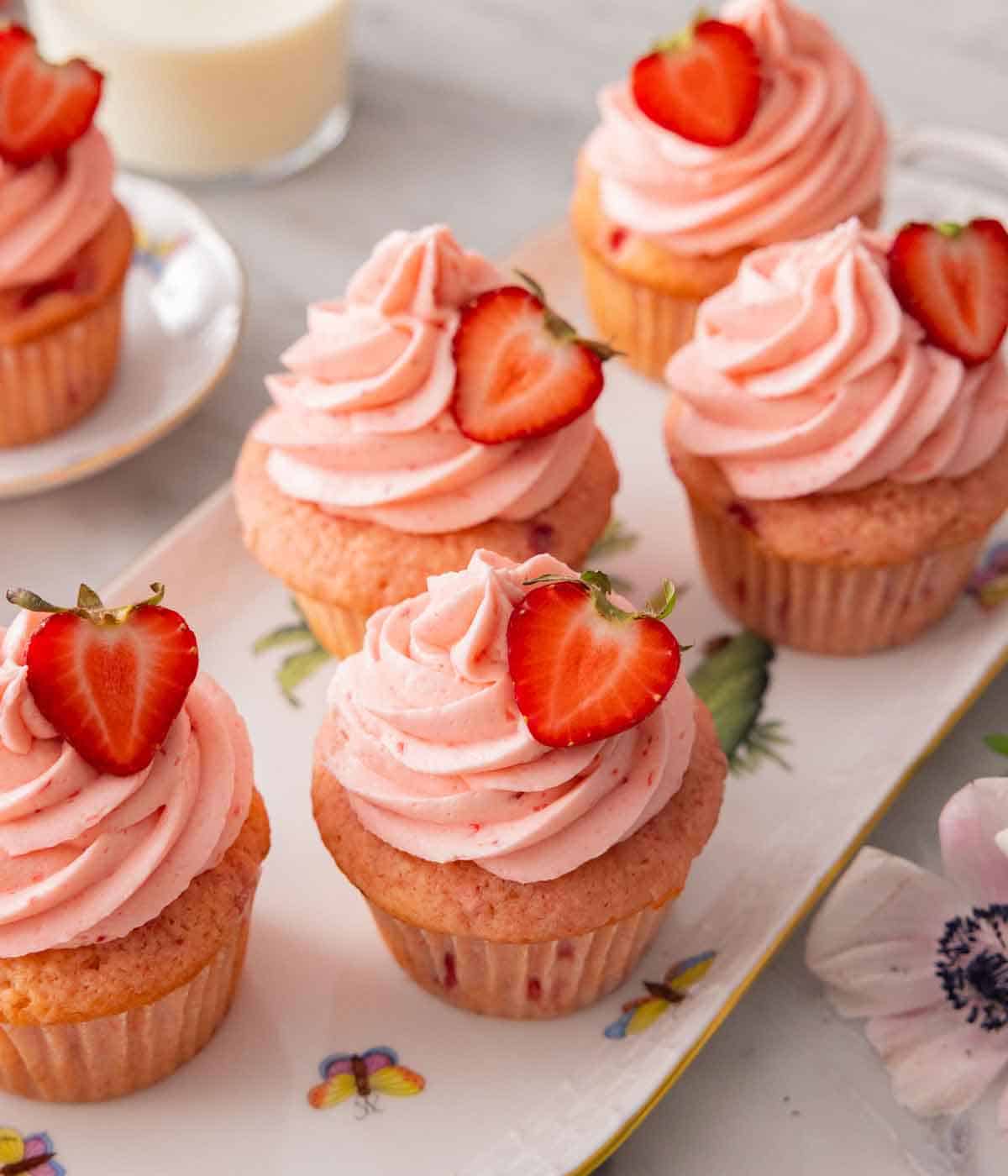 A long platter with multiple strawberry cupcakes with pink frosting and sliced strawberries on top.