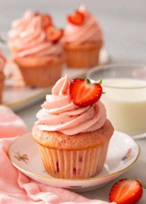 A strawberry cupcake with half a strawberry on top of the pink frosting. More cupcakes and milk in the background.